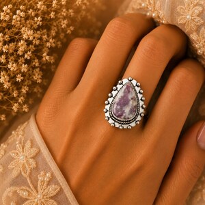 May include: A silver ring featuring a teardrop-shaped purple and white stone set in a decorative metal frame with a beaded edge and a twisted rope detail surrounding the central gemstone, worn on a finger with a visible band design.