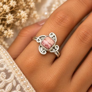 Puede incluir: Un anillo de plata con una piedra preciosa de rodocrosita de corte cuadrado con bandas rosas y blancas, engastada en un marco de metal decorativo con patrones de volutas en los lados, mostrando un acabado pulido y un trabajo de metal detallado.