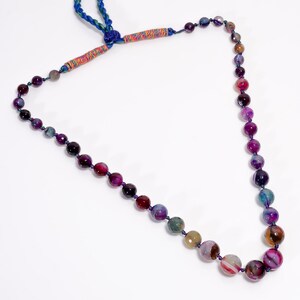 May include: A necklace made of colourful, round gemstones in shades of purple, pink, green, and brown. The necklace has a blue cord with a decorative knot and wrapped accents. The beads vary in size and colour, creating a unique and eye-catching design.