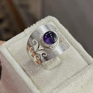 May include: A silver ring featuring a large, round, deep purple gemstone. The ring has a wide band with an ornate, swirling design. The gemstone is set in a textured bezel.