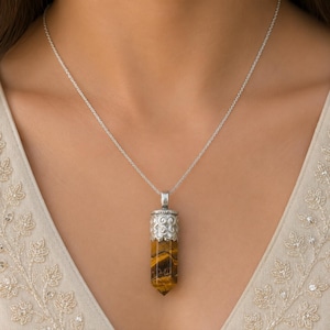 May include: A silver chain necklace featuring a cylindrical tiger's eye pendant. The pendant is capped with a decorative silver metal top that has a swirling, embossed pattern. The stone displays characteristic golden-brown bands.