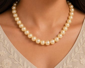May include: A single strand necklace composed of uniform, round, cream-coloured pearls. The pearls feature a smooth, lustrous finish and are strung together in a continuous circular arrangement that rests around the base of the neck.