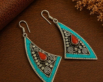 May include: A pair of triangular drop earrings featuring silver metal frames with intricate scrollwork and dotted patterns. Each earring is inlaid with bright turquoise stone accents along the edges and a central teardrop-shaped red coral stone.