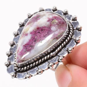 Peut inclure: Bague en argent ornée d'une grosse pierre en forme de goutte d'eau. La pierre est un mélange de blanc et de rose foncé, avec un aspect texturé. Le sertissage de la bague présente une bordure décorative avec de petits accents ronds.