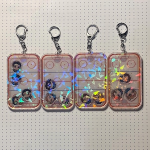 Könnte beinhalten: Vier transparente Acryl-Schlüsselanhänger mit silbernen Schlüsselringen. Jeder Schlüsselanhänger zeigt ein Hockeyfeld-Design mit Charakterillustrationen und schillerndem Glitzer. Die Schlüsselanhänger haben einen roten Rand und sind rechteckig.