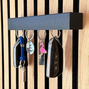 Könnte beinhalten: Ein schwarzer Schlüsselhalter aus Metall mit fünf daran befestigten Schlüsseln. Die Schlüssel haben verschiedene Anhänger, darunter einen blauen und weißen Hausanhänger, einen rosa Flamingo-Anhänger und einen silbernen Anhänger mit dem Text "Unser Zuhause".