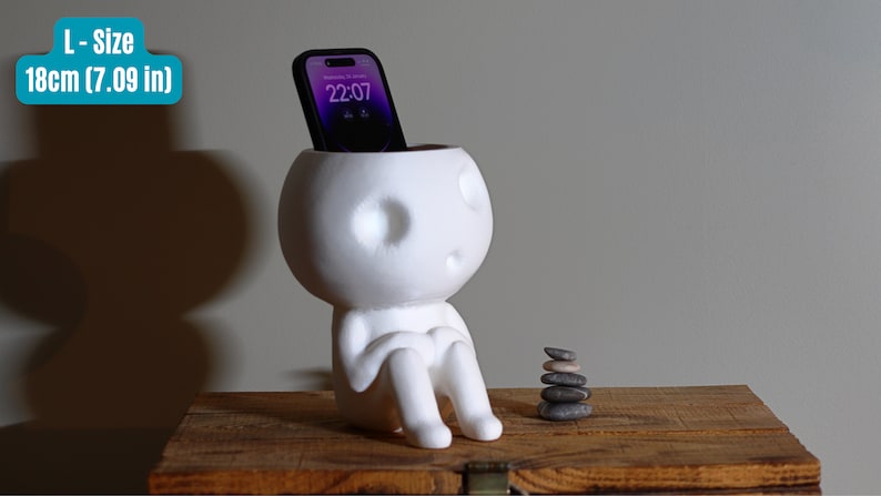 May include: White ceramic planter shaped like a person sitting with a smartphone in its head. The planter is 18 inches tall.  A stack of gray stones is on a wooden surface.
