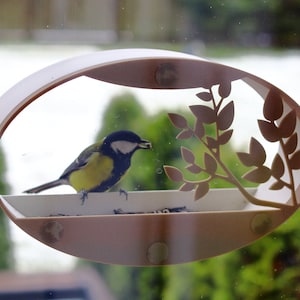 Könnte beinhalten: Ein kleiner blauer und gelber Vogel sitzt auf einem weißen Vogelhäuschen mit einem dekorativen Blattdesign. Der Futterspender ist an einem Fenster befestigt.