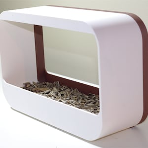May include: A contemporary white bird feeder with a rectangular shape and curved edges. It features a brown trim and is filled with seeds. The text "WHITE / CORK" is visible in the top left corner.