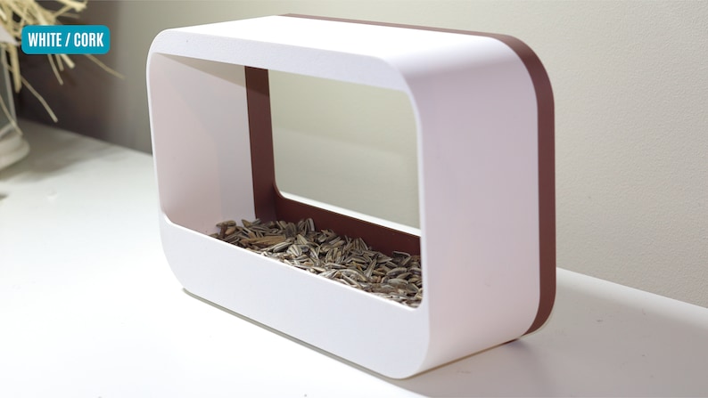 May include: A contemporary white bird feeder with a rectangular shape and curved edges. It features a brown trim and is filled with seeds. The text "WHITE / CORK" is visible in the top left corner.