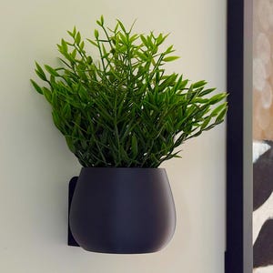 May include: A dark gray, wall-mounted planter filled with vibrant green artificial foliage. The planter has a rounded shape and is mounted on a white wall. A framed zebra print is partially visible in the background.