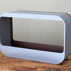May include: A rectangular, open-frame object with rounded corners. The exterior is a light grey, while the interior has a dark brown colour. The object sits on a wooden surface. The text "GREY / CORK" is in a teal rectangle in the upper left corner.