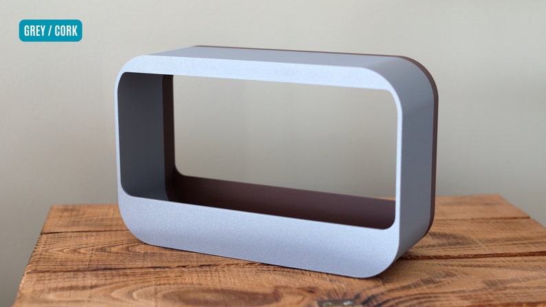 May include: A rectangular, open-frame object with rounded corners. The exterior is a light grey, while the interior has a dark brown colour. The object sits on a wooden surface. The text "GREY / CORK" is in a teal rectangle in the upper left corner.