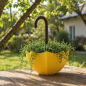 May include: A yellow umbrella-shaped planter filled with green plants. The planter has a dark brown handle and sits on a wooden surface. The background features a green lawn and trees.
