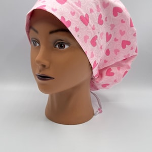 Op de afbeelding: Een roze scrubcap met een patroon van roze harten en krullen. De cap is op een mannequinhoofd. De cap is gemaakt van stof en heeft een strik aan de achterkant.