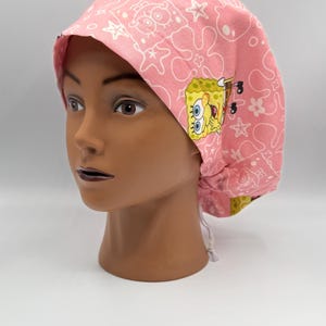 May include: Pink scrub cap featuring a repeating white pattern and a SpongeBob SquarePants graphic. The cap is made of fabric and has a drawstring closure. The cap is designed to be worn in a medical or surgical setting.