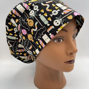 May include: Black scrub hat with a Halloween-themed print featuring candy corn, bats, ghosts, and pumpkins in pink, yellow, and green on a black background.