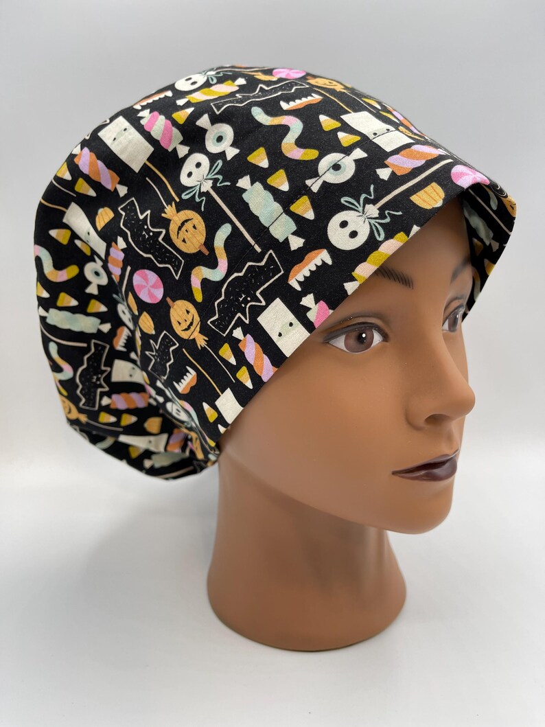 May include: Black scrub hat with a Halloween-themed print featuring candy corn, bats, ghosts, and pumpkins in pink, yellow, and green on a black background.