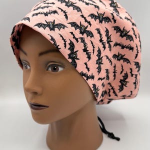 May include: A pink scrub cap with a repeating pattern of black bats. The cap has a black drawstring tie at the back. The cap is displayed on a mannequin head.