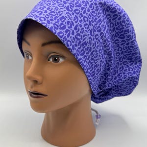 May include: A purple scrub cap with a leopard print pattern. The cap is made of fabric and is designed to be worn in a medical or professional setting. The cap is on a mannequin head.
