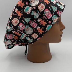 May include: A black scrub cap with a festive Christmas pattern. The cap features Santa Claus, gingerbread men, medical symbols, and other holiday-themed illustrations in pink, teal, and white. The cap is tied with a black drawstring.