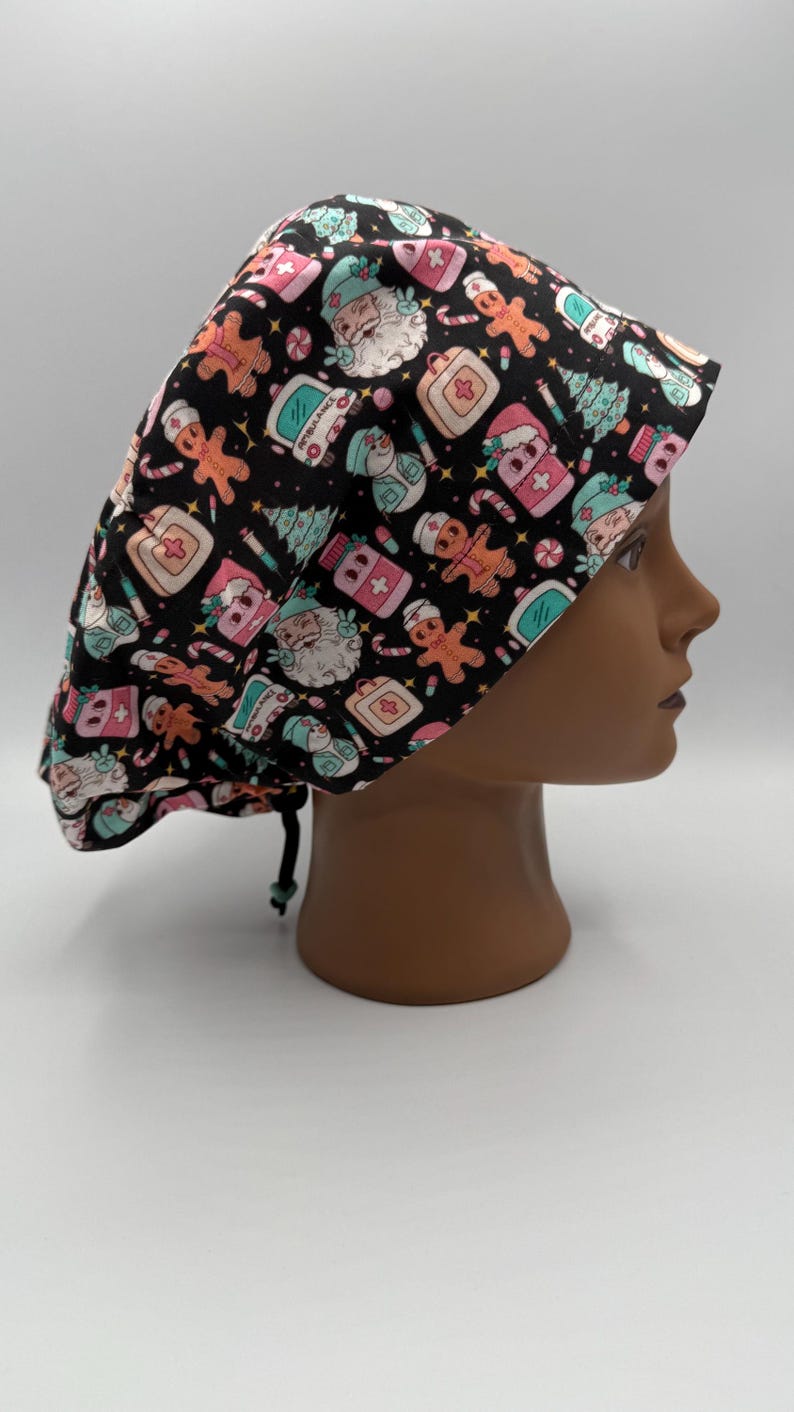 May include: A black scrub cap with a festive Christmas pattern. The cap features Santa Claus, gingerbread men, medical symbols, and other holiday-themed illustrations in pink, teal, and white. The cap is tied with a black drawstring.