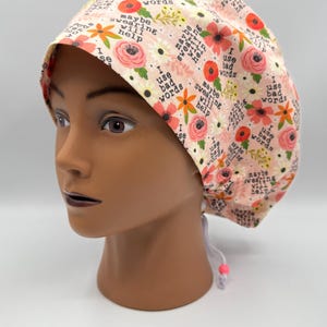 May include: A light pink scrub cap with a floral pattern and the phrases "I use bad words" and "Maybe swearing will help." The cap has a drawstring closure and is displayed on a mannequin head.