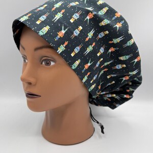 May include: A black surgical scrub cap with a repeating pattern of colorful toy soldier figures. The cap is tied at the back with a black drawstring. The cap is displayed on a mannequin head.