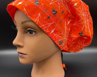 May include: Orange scrub hat with a black spider web and spider pattern. The hat has a drawstring tie.