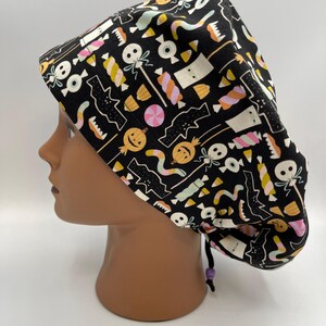 May include: Black scrub hat with a Halloween-themed print featuring candy, pumpkins, bats, and ghosts. The hat has a purple drawstring.