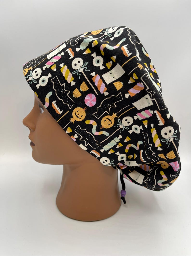 May include: Black scrub hat with a Halloween-themed print featuring candy, pumpkins, bats, and ghosts. The hat has a purple drawstring.
