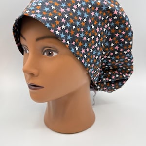 May include: A dark gray scrub cap with a repeating pattern of orange, pink, blue, and white stars. The cap is displayed on a mannequin head against a white background. The cap is designed to cover the hair.