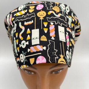 May include: A black scrub hat with a Halloween-themed print featuring candy corn, bats, pumpkins, and other spooky treats.