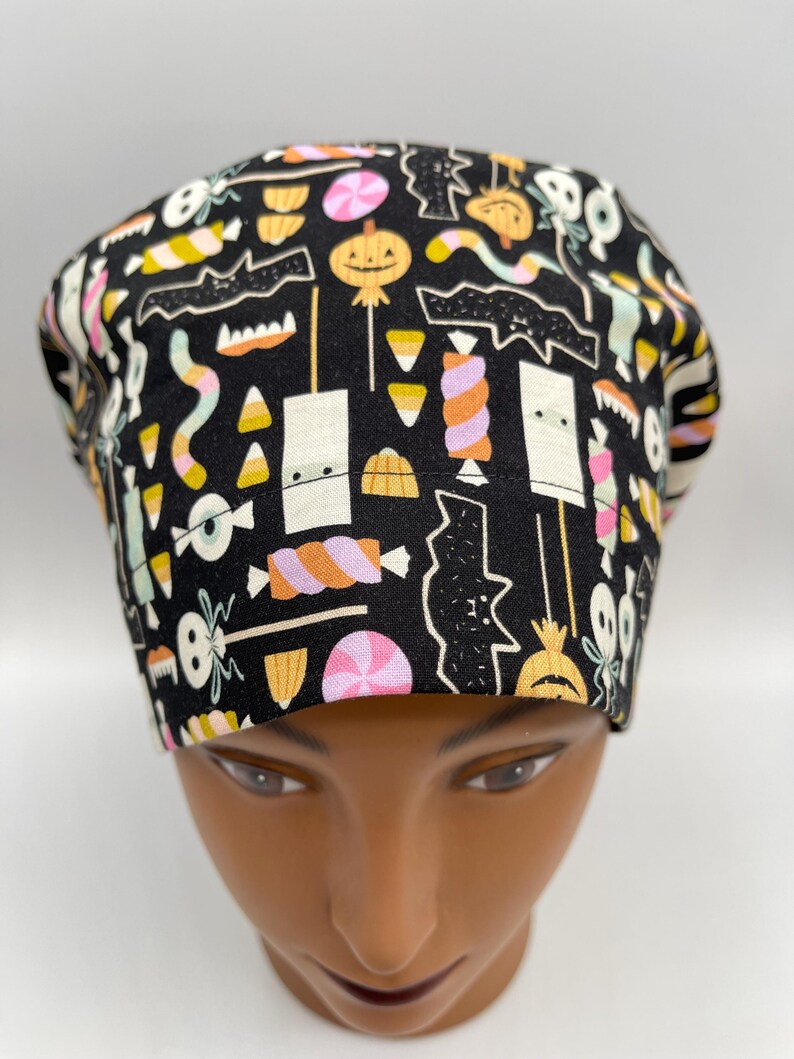 May include: A black scrub hat with a Halloween-themed print featuring candy corn, bats, pumpkins, and other spooky treats.