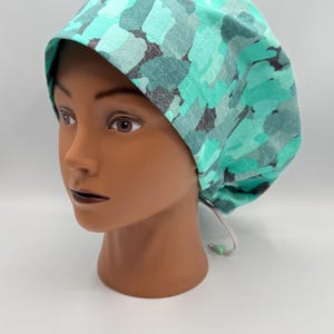 May include: A teal and gray patterned scrub cap. The cap is made of fabric and has a drawstring closure. The cap is on a mannequin head.
