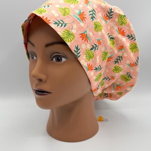 May include: A peach-colored scrub cap with a tropical floral pattern featuring green leaves, orange flowers, and white accents. The cap is displayed on a mannequin head against a white background. The cap has a drawstring.