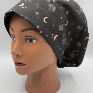 May include: A dark grey scrub cap featuring a pink celestial design of moons, stars, and fireworks. The cap is designed to cover the head and hair, often used in medical or culinary environments.