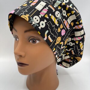 May include: Black scrub hat with a Halloween candy and spooky themed print. The print features candy corn, lollipops, eyeballs, and ghosts.