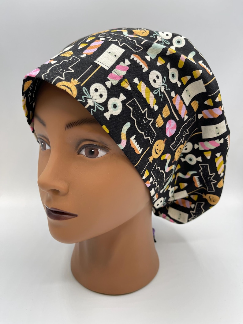 May include: Black scrub hat with a Halloween candy and spooky themed print. The print features candy corn, lollipops, eyeballs, and ghosts.