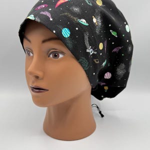 May include: A black scrub cap featuring a colorful space-themed print with planets, stars, rockets, and flying saucers. The fabric has a dark background with scattered white dots and a drawstring cord at the back for an adjustable fit.