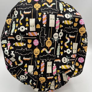 May include: Black fabric scrub hat with a Halloween-themed print of candy, bats, and ghosts. The print is in shades of pink, orange, yellow, and green.
