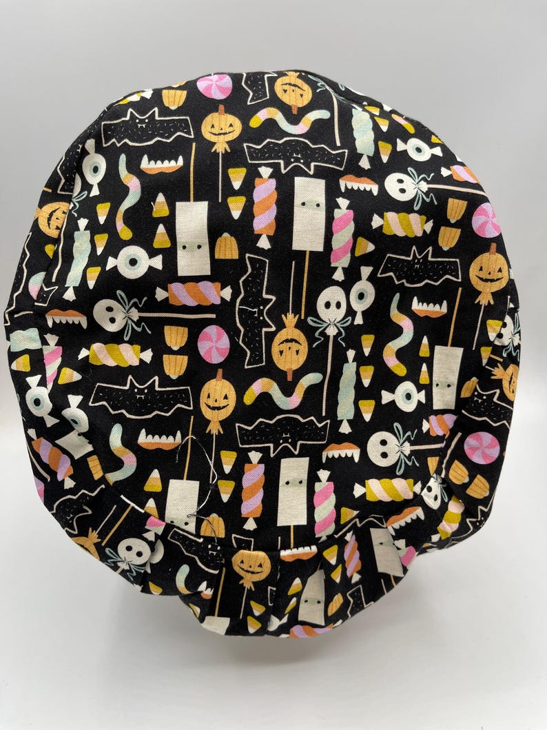 May include: Black fabric scrub hat with a Halloween-themed print of candy, bats, and ghosts. The print is in shades of pink, orange, yellow, and green.