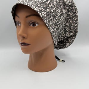 Puede incluir: Un gorro quirúrgico estampado en tonos grises, blancos y rosas, exhibido en una cabeza de maniquí. El gorro presenta un diseño repetitivo en forma de hoja. El gorro tiene un cordón negro en la parte posterior para ajustarlo. El fondo es blanco liso.