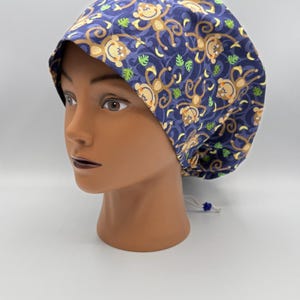 May include: A blue scrub cap with a repeating pattern of cartoon monkeys, bananas, and leaves. The cap is made of fabric and has a drawstring closure at the back. The cap is displayed on a mannequin head.