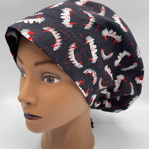 May include: Black surgical scrub cap with a repeating pattern of white fangs and red blood splatters. The cap is made of a soft fabric and is designed to cover the head and hair. The cap is tied in the back.