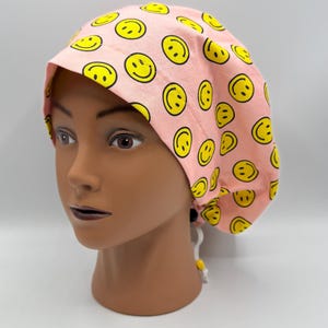 Może przedstawiać: Różowy czepek chirurgiczny z powtarzającym się wzorem żółtych uśmiechniętych buziek. Czepek jest wykonany z tkaniny i ma wiązanie z tyłu do regulacji. Czepek jest przeznaczony do noszenia w środowisku medycznym lub zawodowym.