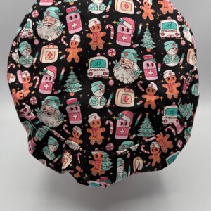 May include: A black scrub cap with a festive Christmas-themed pattern. The design includes gingerbread men, Santa Claus, Christmas trees, ambulances, and medical symbols in pink, white, and teal. The cap is designed for medical professionals.