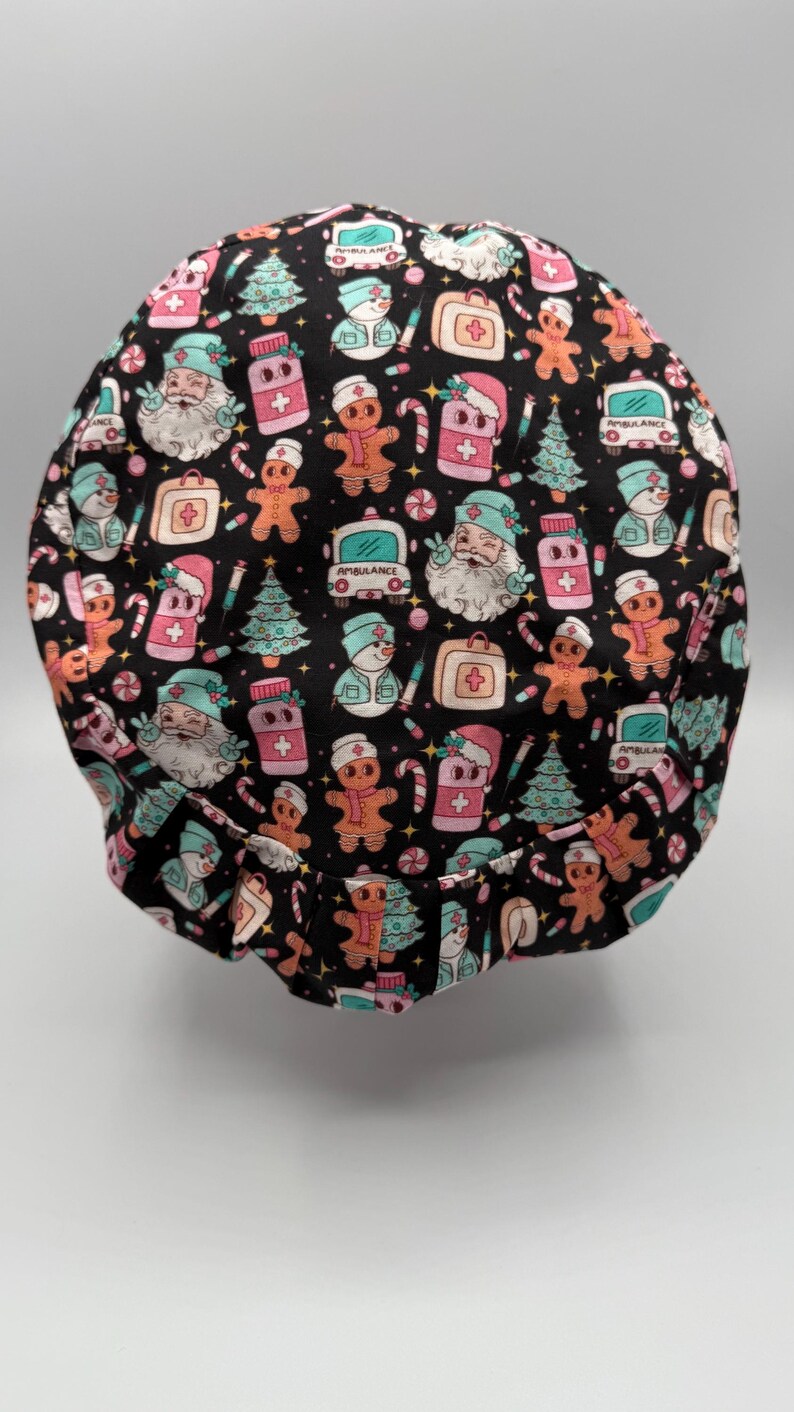 May include: A black scrub cap with a festive Christmas-themed pattern. The design includes gingerbread men, Santa Claus, Christmas trees, ambulances, and medical symbols in pink, white, and teal. The cap is designed for medical professionals.