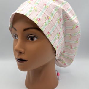 Può includere: Un cappuccio per scrub a fantasia floreale rosa e bianco. Il cappuccio presenta un motivo ripetuto di rose rosa e foglie verdi su uno sfondo rosa chiaro con strisce verticali. Il cappuccio è progettato per essere indossato in un ambiente medico o sanitario.