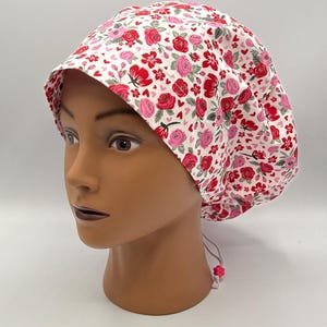 Könnte beinhalten: Eine weiße OP-Haube mit einem Blumenmuster aus roten und rosa Rosen und kleinen rosa Herzen. Die Haube hat einen Schirm und einen verstellbaren Kordelzug. Die Haube befindet sich auf einem Schaufensterkopf.